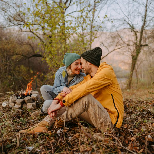 Photo of young couple spending autumn day in nature, away from the city. They are sitting next to a campfire to get warm.