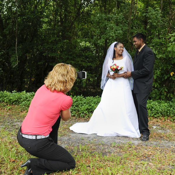 Bride and groom posing for wedding portraits near a woodland while their wedding photographer crouches down to take a photo