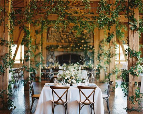 An indoor wedding reception area with greenery, white tables, floral centerpieces, and wooden chairs.