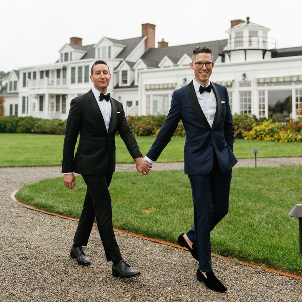 Groom in Black Tuxedo Holding Hands With Groom in Dark Navy Tuxedo Walking in Front of White Mansion