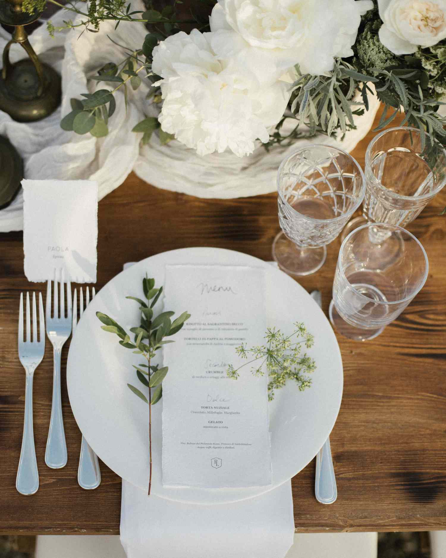 Wedding reception tablescape with wood tables, white runner, white florals, white plates, and greenery sprigs