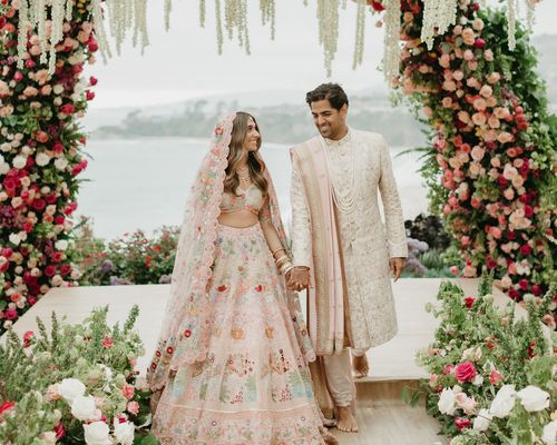 Bride in Pink and Floral Traditional Indian Dress Holding Hands With Groom in Cream Kurta with Light Pink Chunni Standing Under Pink and White Flower Arches