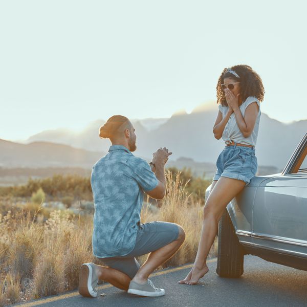 Man on One Knee Holding an Engagement Ring Box and Woman Looking Surprised as She Rests on a Car Door