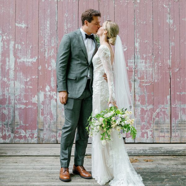 Portrait of Groom in a Gray Suit and Bride in Lace Gown Holding a Loose Bouquet Against Rustic Red Wall