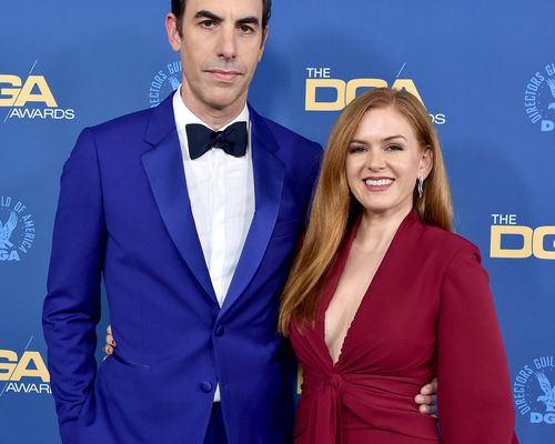 Sacha Baron Cohen wearing a blue suit and bow-tie posing next to Isla Fisher wearing a cranberry red dress on the DGA Awards red carpet