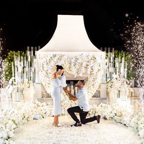 groom on one knee proposing to bride dressed in white and standing in front of fireplace decorate with heart-shaped floral arrangement and candles