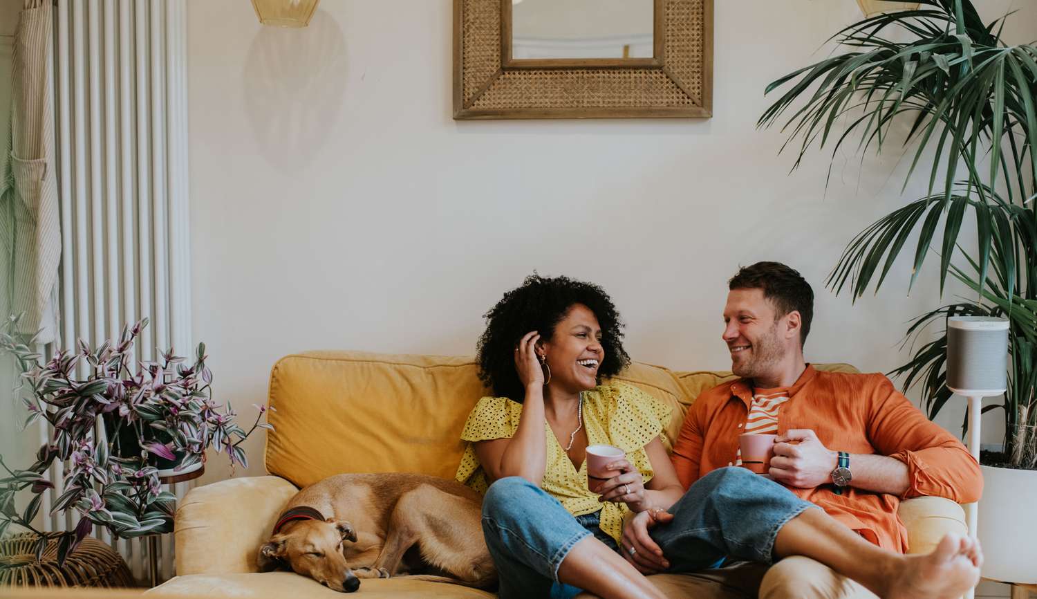 Woman and Man Facing Each Other and Smiling While Sitting on the Couch and Holding Coffee Mugs Next to Their Dog