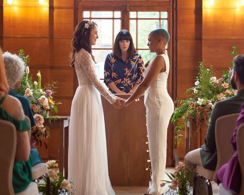 Two brides in white wedding attire exchange vows during a wedding ceremony officiated by a civil officiant. 