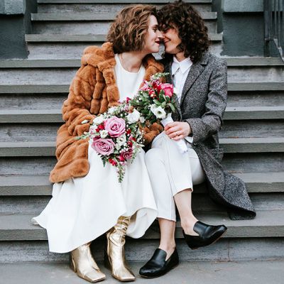 Two brides pose on a staircase with winter wedding bouquets featuring greenery, white flowers, and pink roses.