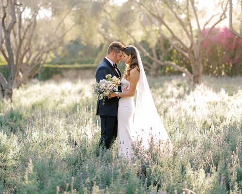 Bride in Wedding Dress and Long Veil Posing With Groom in Navy Tuxedo In Field With Tall Grass