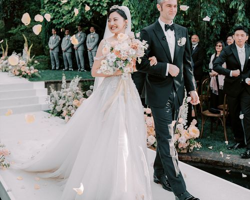 Bride in White Wedding Dress and Veil Holding Flower Decor Walking Next to Groom in Black Tuxedo Down Aisle 