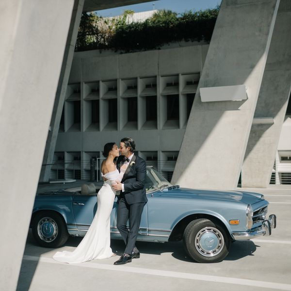 Bride in Strapless Wedding Dress Kissing Groom in Black Tuxedo in Front of Vintage Blue Car Parked in Parking Garage