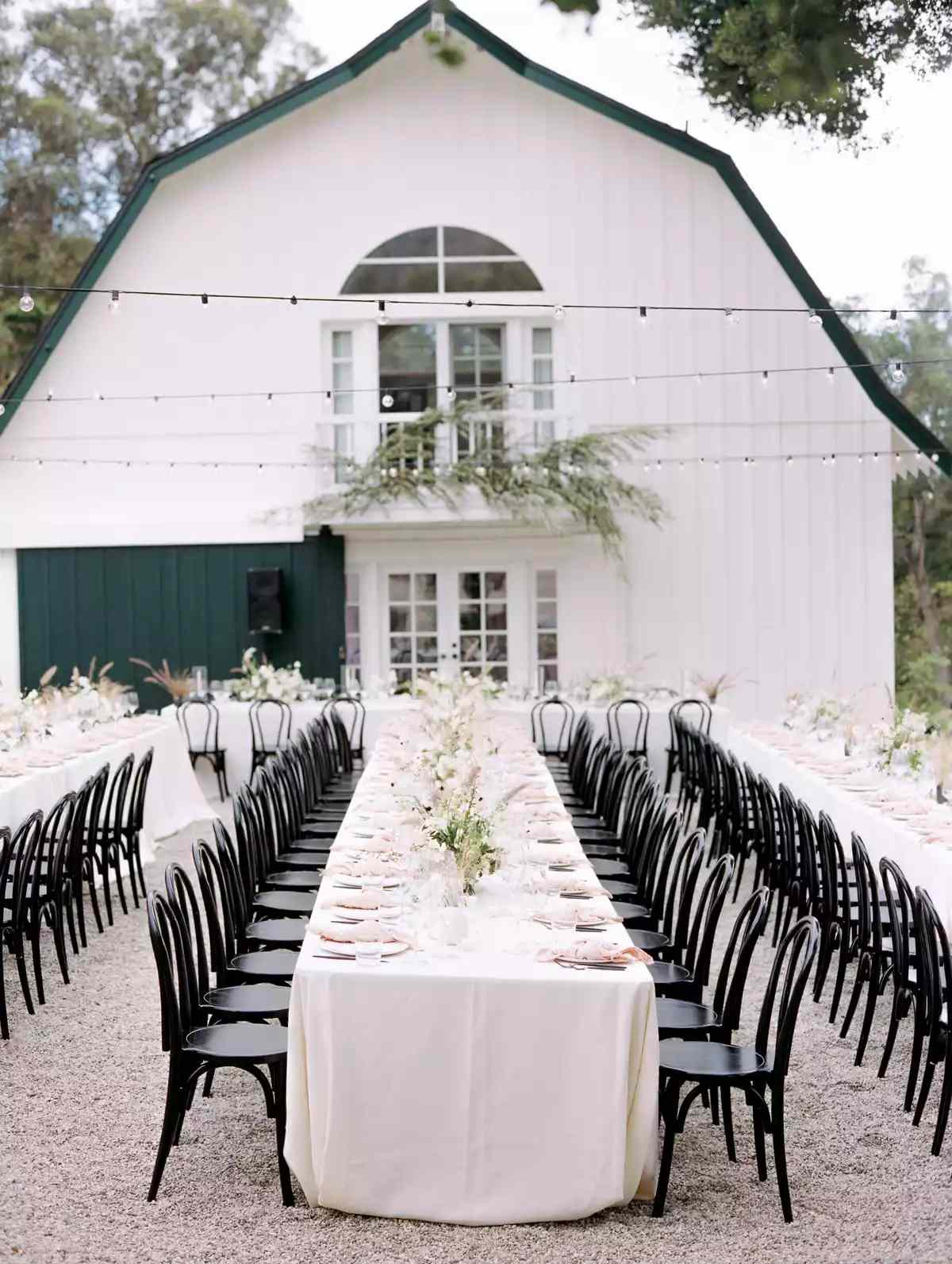 Long Wedding Reception Tables at Rustic Outdoor Venue with Black Chairs, Neutral Linens, and Floral Arrangements