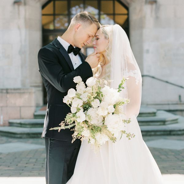 Bride In Wedding Dress and Veil Holding Large White Bouquet Posing With Groom in Black Tuxedo