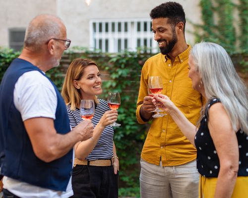 Wife in striped shirt and husband in yellow shirt holding glasses of rose wine with in-laws in their back yard.