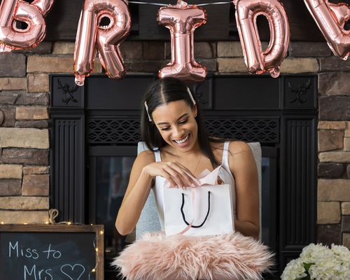 Bride-to-be at a bridal shower smiling while opening a bridal shower gift surrounded by a pink balloon sign and white flowers