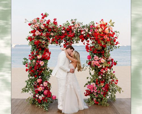 Bride and Groom Kissing Beneath Pink and Red Floral Ceremony Altar at Beachfront Wedding Ceremony