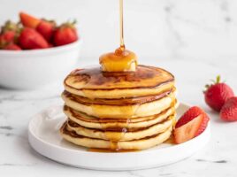 Side view of a stack of pancakes with butter on top and syrup being poured over the top.