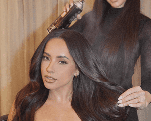 A woman sitting in a chair while a hairstylist sprays her long brown hair with hairspray