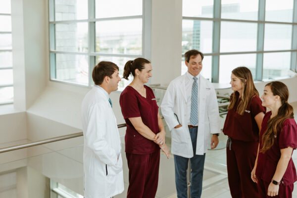 A CIS nurse in burgundy scrubs works at a computer stations while 2 more nurses review a patient chart in the background