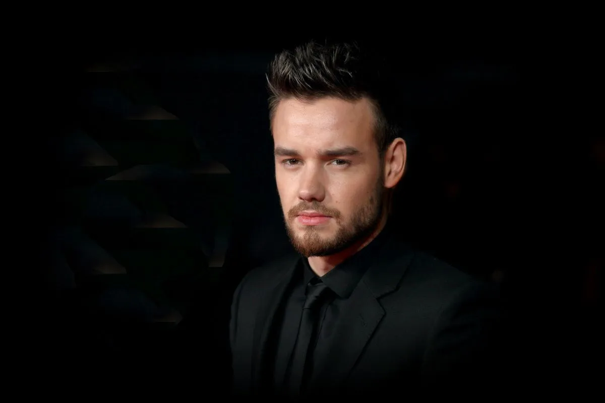 Liam Payne wears a black shirt and stands against a black background.