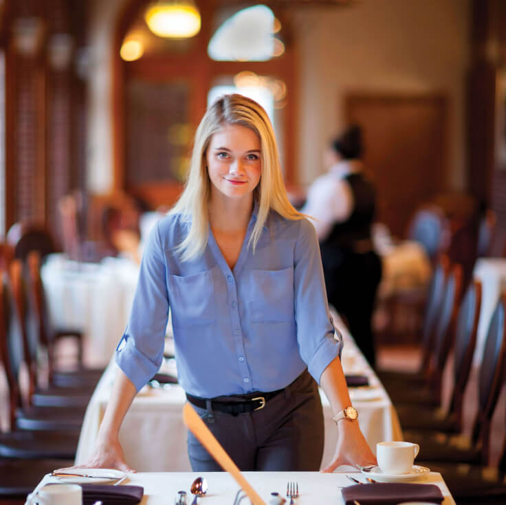 A woman with long blonde hair, wearing a blue blouse and dark trousers, stands leaning forward on a table in a restaurant. The table is set with cups and plates, hinting at her role in food service management. In the background, another person in uniform is seen walking away.