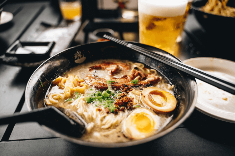 A bowl of ramen noodle soup is garnished with sliced soft-boiled eggs, green onions, and pieces of pork. Black chopsticks rest on the edge of the bowl, and a frothy glass of beer is in the background. Other dishes are slightly blurred in the background.