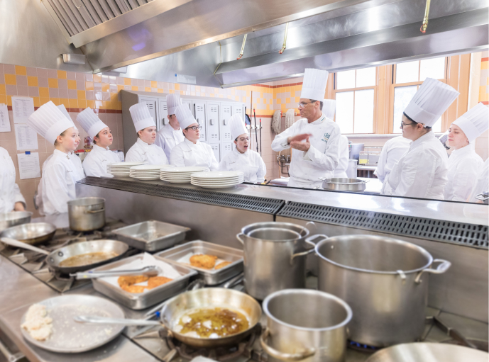 A group of culinary students in white chef uniforms and tall hats gather around an instructor in a commercial kitchen. The instructor appears to be giving a demonstration, and various pots, pans, and plates are visible on the stovetop and counters.