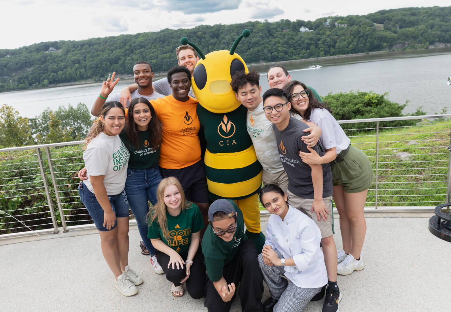 A group of eleven diverse students smiling and hugging a large bee mascot dressed in a yellow and black costume with "CIA" on its chest. They are standing on an outdoor terrace with a river and green hills in the background.