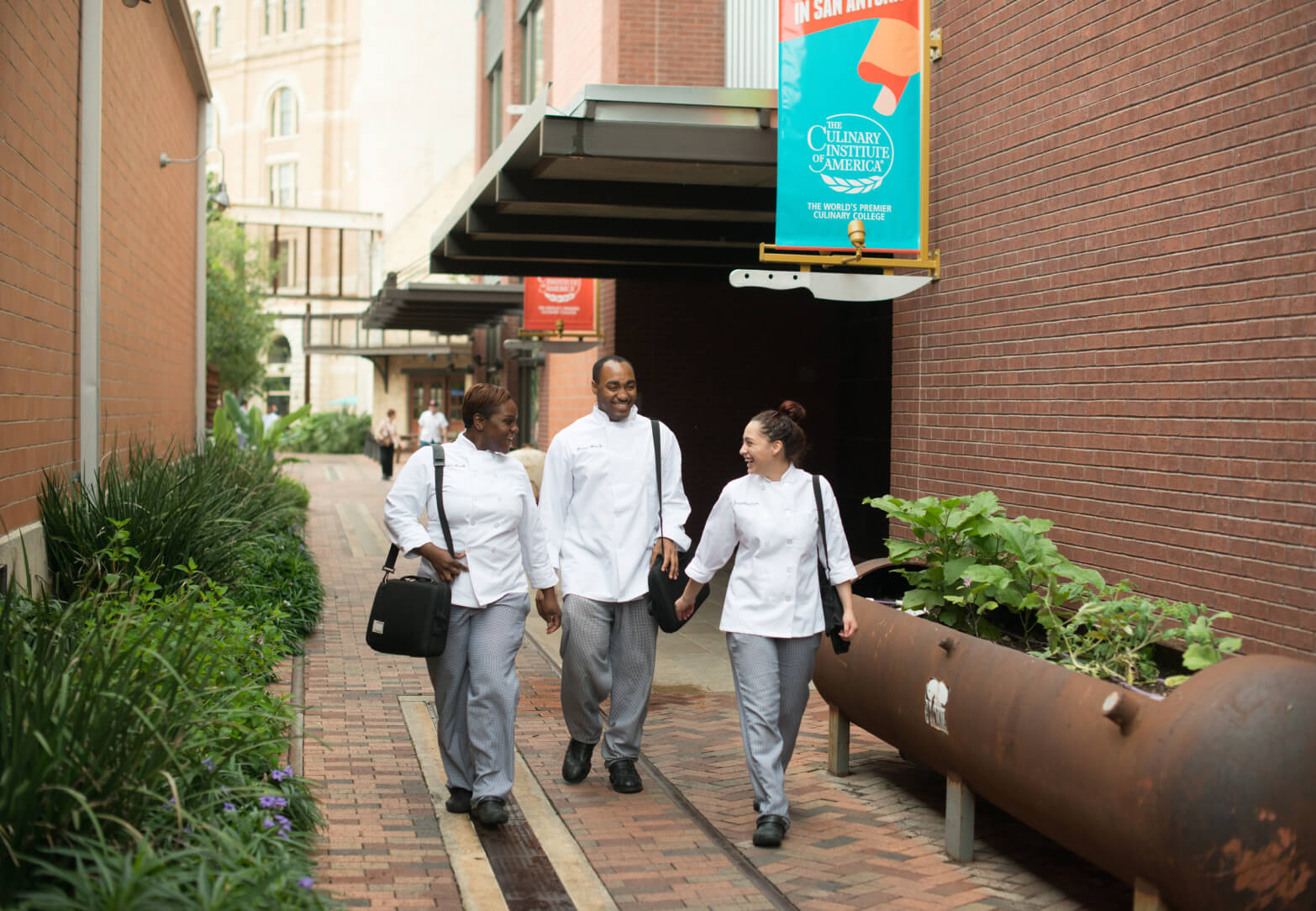 Three people in chef uniforms walk together down a brick pathway between buildings. They are talking and smiling. One person carries a black bag. There is a sign overhead that reads "The Culinary Institute of America." Greenery and plants are on one side of the walkway.