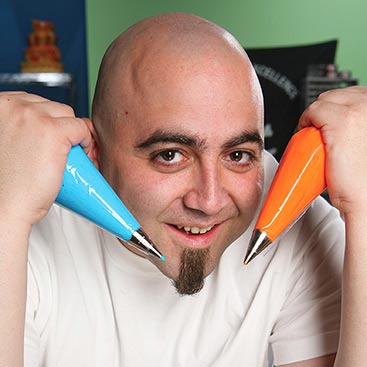 A bald man with a goatee, wearing a white shirt, holds two piping bags with different colored icing, one blue and one orange, near his face. He smiles as he poses playfully in a brightly lit room with colorful backgrounds.