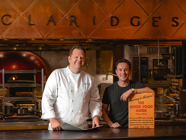 Two men are standing in a professional kitchen under a sign that reads 