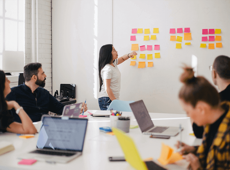 Pessoas apontado para vários post-it organizados em uma parede, à frente de uma mesa com pessoas reunidas.