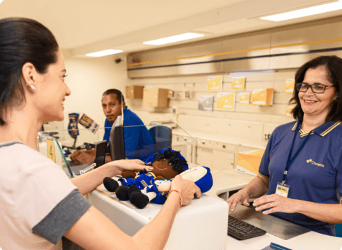 Mulher em frente ao balcão sendo atendida por outra mulher, funcionária dos Correios, que recebe uma encomenda em mãos. 