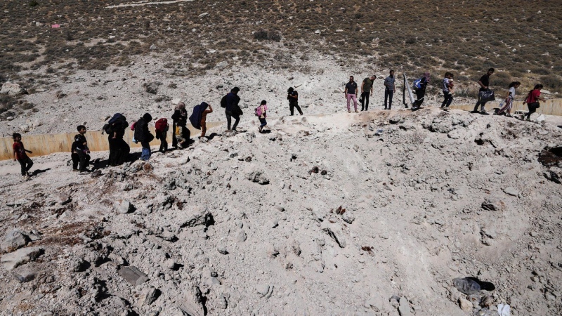 People carry their luggage as they cross into Syria on foot, through a crater caused by Israeli airstrikes aiming to block Beirut-Damascus highway at the Masnaa crossing, in the eastern Bekaa Valley, Lebanon, Saturday, Oct. 5, 2024. (AP Photo/Hassan Ammar)