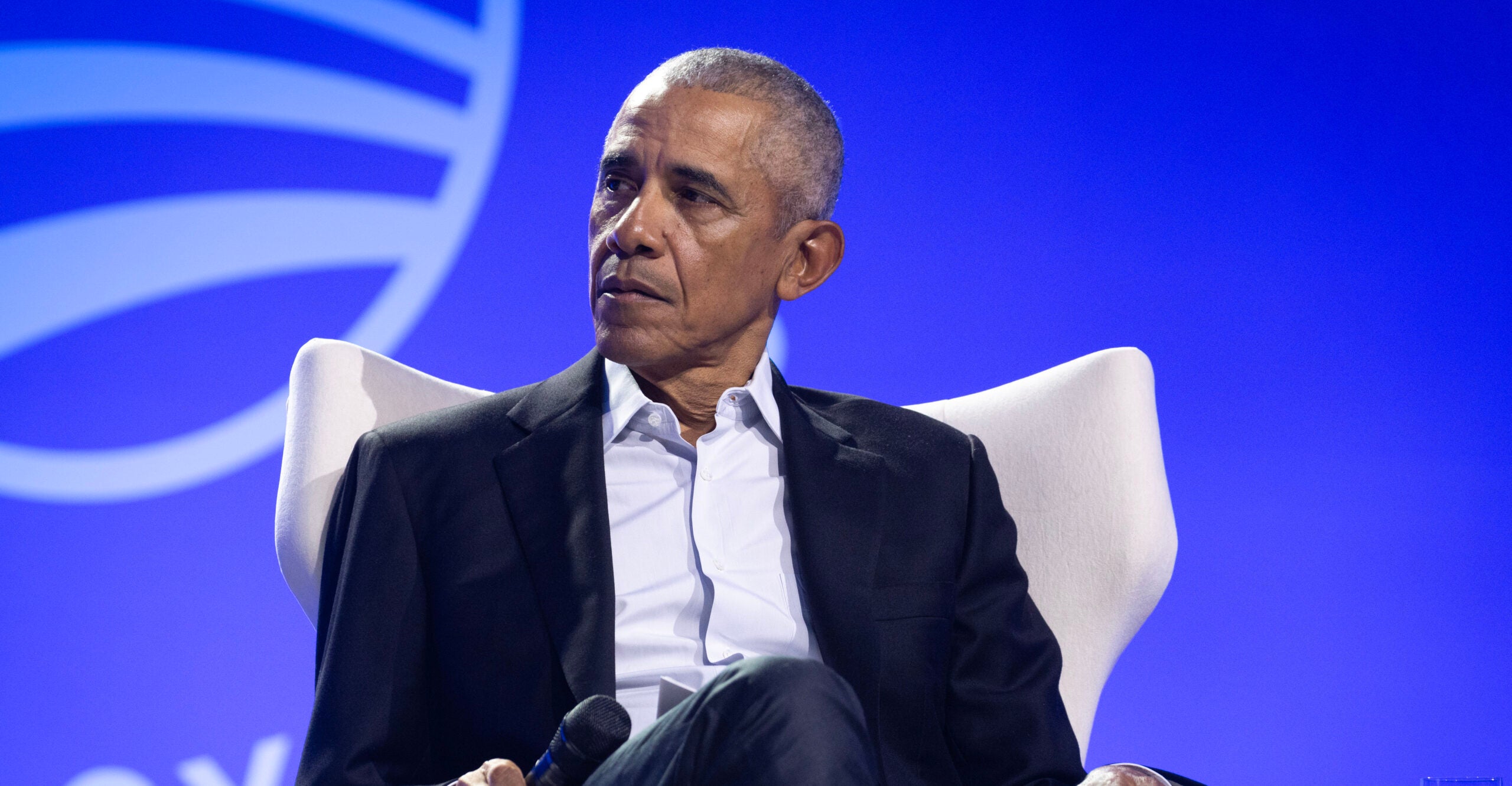 Barack Obama, seated, wears a dark blue suit and white dress shirt as he holds a microphone in his right hand.