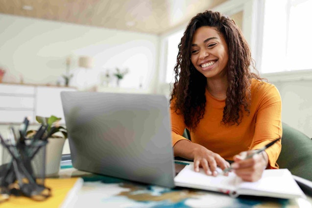 A woman smiles and takes notes while she looks at a screen.