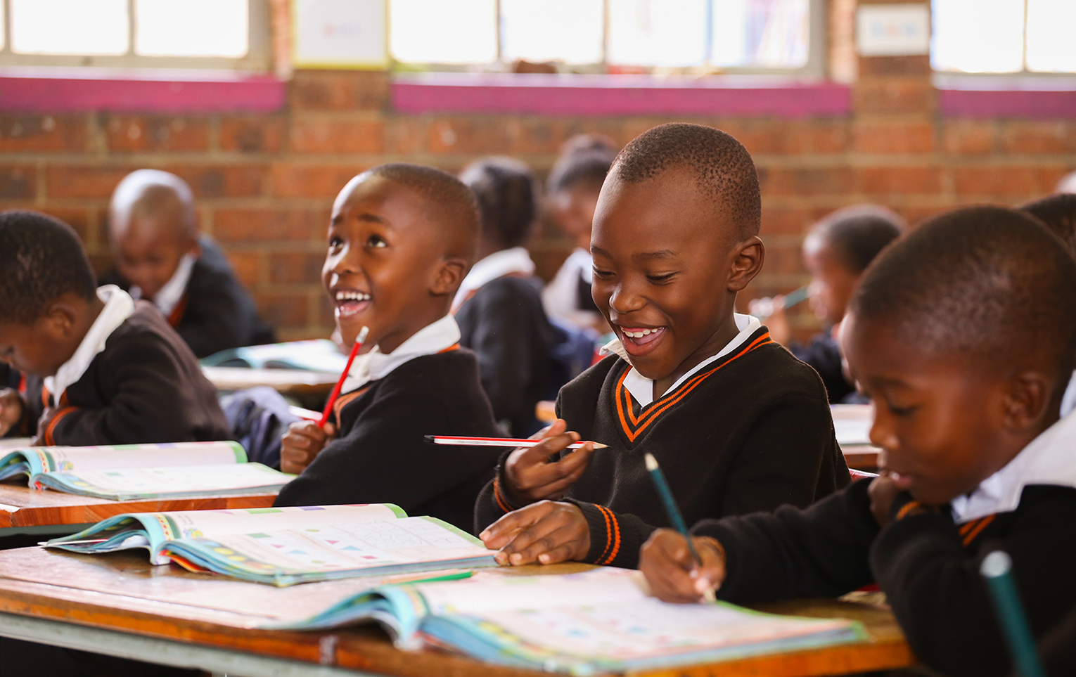 Three boys smile while completing schoolwork in a classroom at Sedibeng Primary School in Thembisa, Johannesburg, South Africa.