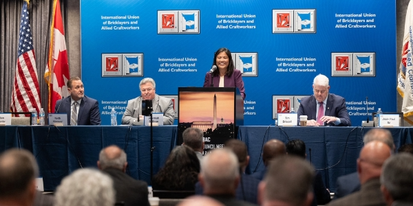 Acting Secretary Su speaks at a podium in front of an International Union of Bricklayers and Allied Craftworkers banner. Several people are seated on either side of her. 