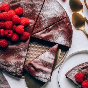 An overhead view of a flourless chocolate cake topped with raspberries and powdered sugar. A slice of cake is resting on a serving utensil.