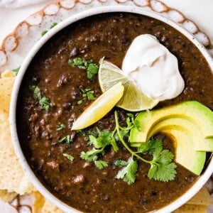 A bowl of black bean soup topped with avocado, cilantro, lime and vegan sour cream served alongside tortilla chips.