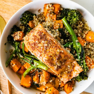 A white serving bowl with tri-colored quinoa and roasted sweet potatoes and broccolini mixed together and topped with mustard maple salmon.