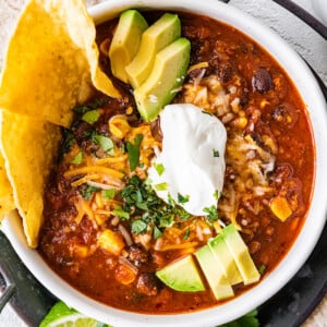 A white bowl full of turkey chili, topped sour cream, shredded cheese, avocado slices, fresh cilantro, and tortilla chips.