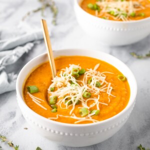 Two bowls of carrot parsnip soup garnished with scallions and shredded gruyere cheese.