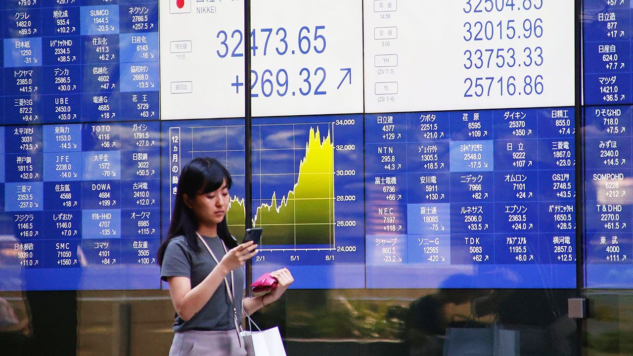 A young woman in Japan checks her phone while standing in front of a screen displaying financial data.