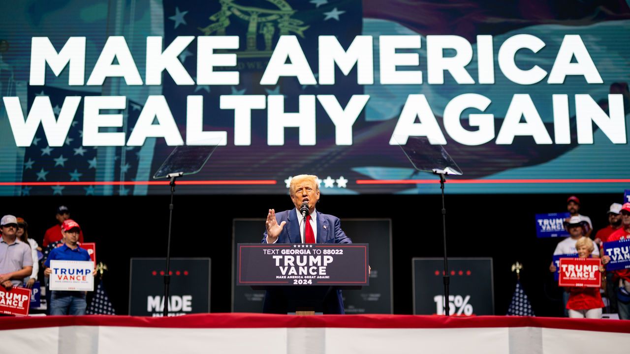 Donald Trump speaks at a campaign rally with the words Make America Wealthy Again on a screen behind him.