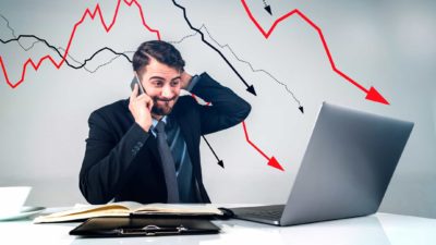 A shocked man sits at his desk looking at his laptop while talking on his mobile phone with declining arrows in the background representing falling ASX 200 shares today