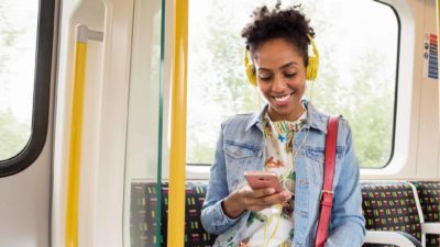 A woman smiles as she sits on the bus using her phone and listening to music through headphones.