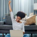 A woman sitting in her lounge room punches the air in a gesture of success, having seen the rising IAG share price on her laptop