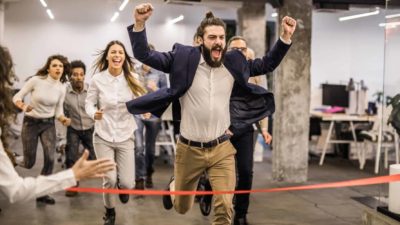 Man in an office celebrates at he crosses a finish line before his colleagues.